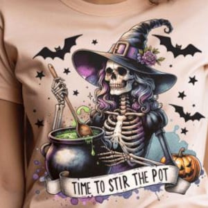 May include: A beige t-shirt featuring a colorful illustration of a skeleton witch stirring a green potion in a cauldron. The witch wears a black hat with a purple brim and is surrounded by bats, stars, and a pumpkin. The banner reads "TIME TO STIR THE POT."