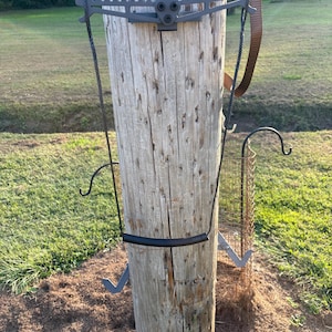 May include: A gray metal climbing system attached to a weathered wooden pole. The system includes a platform, steps, and a safety strap. The pole is surrounded by grass and mulch, with a blurred background of trees and sky.