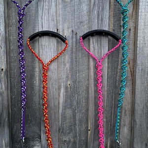 May include: Four colorful dog leashes with black handles are displayed against a weathered wooden background. The leashes are purple, red, pink, and teal, each with a unique woven pattern. The handles are curved and attached to the wall.