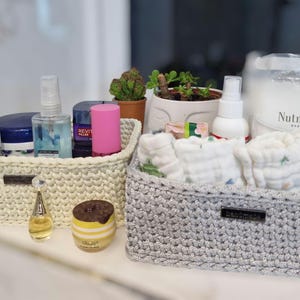 May include: Two handmade crochet baskets, one cream and one grey, filled with toiletries, baby items, and a small potted plant. A jar labelled "Nutmeg" is visible. The baskets are on a white surface.