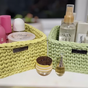 May include: Two rectangular crochet baskets in light yellow and sage green, each featuring a small dark rectangular brand tag on the front. The baskets are filled with various personal care items, including pink bottles, a clear spray bottle, and jars.