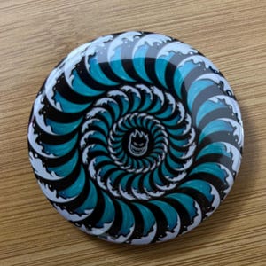 May include: A round button with a black and white spiral design featuring a blue wave pattern. The center of the spiral has a small black and white graphic of a skull with a flame.