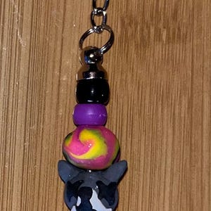 May include: A silver keychain with a black, white, and purple beaded charm. The charm features a swirl pattern and a black and white animal head.