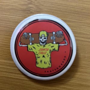 May include: A round button with a red background featuring a skeleton wearing a yellow shirt and hat, holding a skateboard. The text "SANTA CRUZ SKATEBOARDS" is printed around the edge of the button.