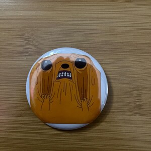 May include: A circular badge with a cartoon character illustration. The character is orange with black eyes and an open mouth, conveying surprise. The badge has a white border and is displayed on a wooden surface.
