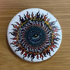 Könnte beinhalten: Ein runder Knopf mit einem bunten Auge, das von Flammen umgeben ist. Das Auge ist blau, grün und schwarz, und die Flammen sind rot, orange, gelb und schwarz.
