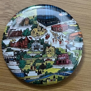 Puede incluir: Un pin redondo y colorido con una ilustración detallada de una ciudad con edificios, árboles y personas. El pin tiene un acabado brillante e incluye texto como "Quality Book Shoppe" y "UNIVAC".