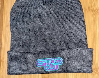 Könnte beinhalten: Graue Strickmütze mit einem gestickten "Spaced Out"-Patch in Lila und Türkis.