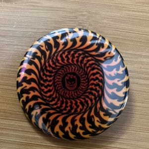 May include: A round button with a black, orange, and yellow swirl pattern. The swirl pattern is reminiscent of flames. The center of the button has a black and red logo.