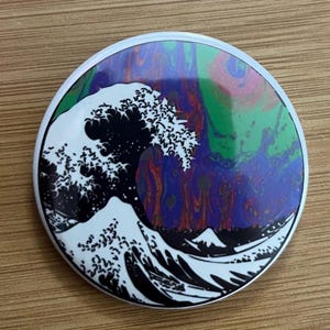 May include: A round, white-bordered button featuring a stylized black and white wave design, reminiscent of Japanese art. The background has a swirling pattern of blue, green, and purple colors.