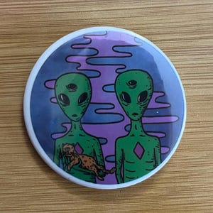 May include: A round button featuring two green aliens with three eyes each, set against a purple and blue background. One alien holds a brown and orange cat. The button has a white border.