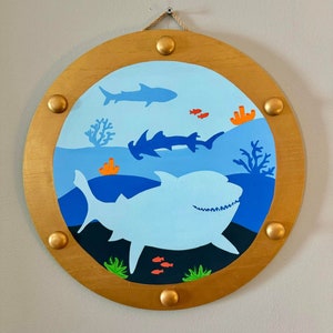 May include: A round wooden wall decoration with a gold frame depicting a blue ocean scene with sharks and fish. The frame has gold accents and a rope hanger.