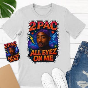 Puede incluir: Camiseta blanca con un diseño gráfico de 2Pac. El diseño incluye el texto "2PAC" y "ALL EYEZ ON ME", un retrato de 2Pac con una bandana morada y un fondo colorido con rayos y corazones.