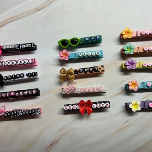 May include: A collection of decorative clothespins with various designs. Some pins feature phrases like "SPARKLE" and "GIRL POWER" spelled out with letter beads. Others have bows, flowers, and the word "ALOHA".