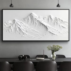 Puede incluir: Escultura en relieve de montañas blancas enmarcada. La obra de arte presenta una cordillera detallada con picos y valles texturizados. El marco negro contrasta con el relieve blanco. La obra se exhibe sobre una mesa de comedor con sillas negras.