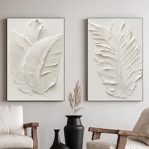 May include: Two framed white feather art prints. The artwork features textured, three-dimensional feather designs against a neutral background. The frames are black, and the overall aesthetic is minimalist and modern, suitable for home decor.