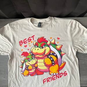 May include: A beige t-shirt with a cartoon image of two green and yellow characters with red accents. The text "BEST FRIENDS" is printed on the shirt.