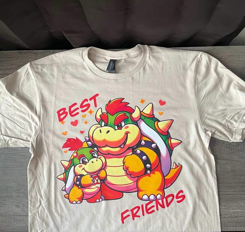 May include: A beige t-shirt with a cartoon image of two green and yellow characters with red accents. The text "BEST FRIENDS" is printed on the shirt.