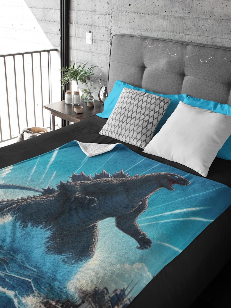 May include: A blue and white fleece blanket featuring a Godzilla graphic. The blanket depicts a Godzilla monster emerging from the water against a blue background with white streaks of light.