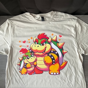 May include: A white t-shirt with a green and yellow cartoon image of two characters from the Super Mario Bros. video game series. The larger character is Bowser, the Koopa King, and the smaller character is a Koopa Troopa. Both characters are smiling and have hearts around them.