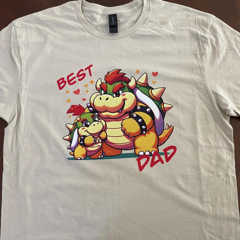 May include: A light beige t-shirt with a graphic print of two cartoon characters. The larger character is a green, yellow, and red Bowser with a spiky red shell. The smaller character is a green, yellow, and red Bowser Jr. with a spiky red shell. The text "BEST" is above the characters and the text "DAD" is below the characters.
