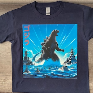 May include: A navy blue t-shirt with a graphic print of Godzilla standing in the ocean with a Japanese word "ゴジラ" in red on the left side of the image.
