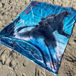 May include: A blue beach towel featuring a Godzilla graphic and the Japanese word "Godzilla" in red. The towel is lying on a sandy beach.