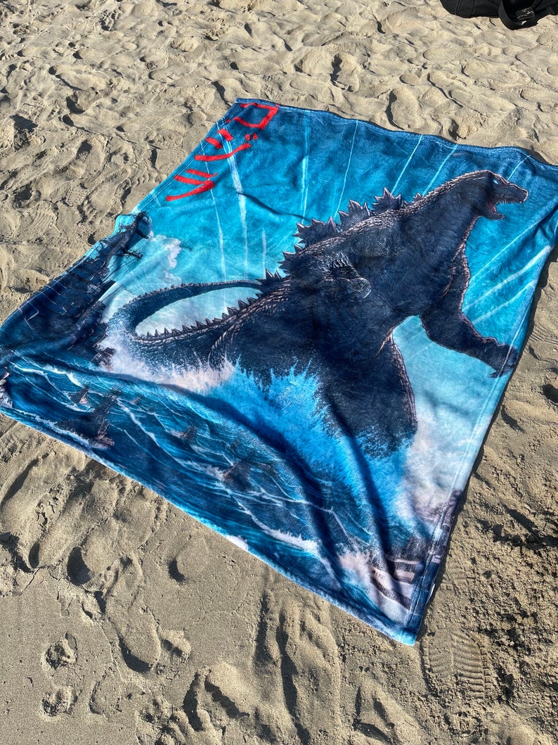 May include: A blue beach towel featuring a Godzilla graphic and the Japanese word "Godzilla" in red. The towel is lying on a sandy beach.