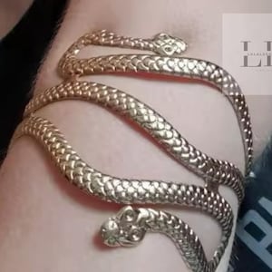 Puede incluir: Pulsera de serpiente dorada con diseño texturizado. La pulsera está hecha de metal y tiene una cabeza de serpiente en cada extremo.