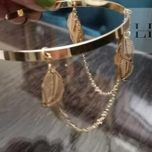 Pode incluir: Pulseira dourada com uma corrente e três pingentes de pena. A pulseira é feita de metal e tem um design simples.
