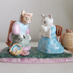 May include: A ceramic figurine set featuring two anthropomorphic cats sitting on chairs on a green rug. One cat is brown and white wearing a pink and blue outfit, while the other is white wearing a blue dress. A small kitten peeks from a wooden barrel.