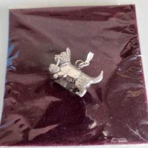 May include: A silver-toned metal charm shaped like a Scottish Terrier dog playing with a ball of yarn. The charm features a small jump ring at the top for attachment and is presented against a dark maroon background inside a clear plastic sleeve.
