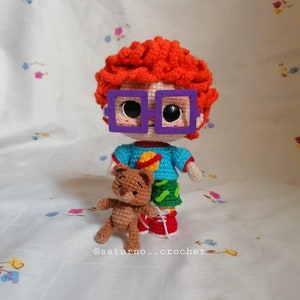 May include: A crocheted doll with red curly hair, purple glasses, a blue and green shirt, and red shoes. The doll is holding a brown crocheted teddy bear. The doll is standing on a white background with a floral pattern.