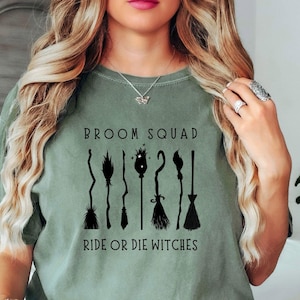 May include: Sage green t-shirt with the text "Broom Squad Ride or Die Witches" in black. The design features stylized black broom illustrations. The shirt is a comfortable, casual style.
