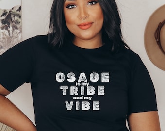 May include: Black t-shirt with the text "OSAGE is my TRIBE and my VIBE" in distressed white lettering. The shirt is a crew neck style and is worn by a person. The person is wearing blue jeans.