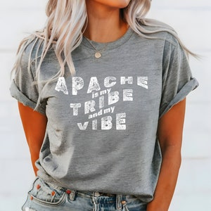 May include: A grey t-shirt featuring the text "APACHE is my TRIBE and my VIBE" in distressed white lettering. The shirt has rolled-up sleeves and a relaxed fit. The model is wearing blue jeans.