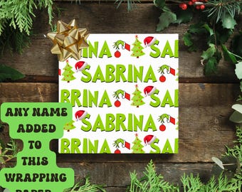 May include: White wrapping paper with a repeating pattern of green text that says "SABRINA", Christmas trees, Grinch hands, and Santa hats. A gold bow is attached to the top left corner. The wrapping paper is surrounded by Christmas greenery.