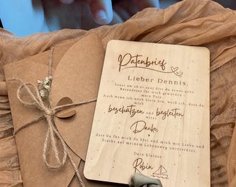 Puede incluir: Una tarjeta rectangular de madera con texto grabado en alemán y un pequeño icono de velero, acompañada de un sobre de papel marrón atado con cordel y una ramita de paniculata seca, con una cinta verde salvia en el borde inferior.