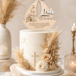 May include: A wooden cake topper shaped like a sailboat featuring the text Zur Taufe and Milo, with an engraved illustration of animals, a rainbow, and a Bible verse. The topper is placed on a white cake decorated with a cross and dried flowers.