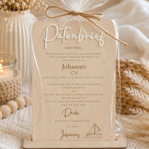 May include: A rectangular light wood plaque with rounded corners, held in a matching wooden base and wrapped in clear plastic with a jute twine bow. The text reads Patenbrief, Lieber Mathis, Johannes, and includes a sailboat graphic.