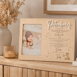 May include: A rectangular wooden frame featuring a photo of a baby wearing a striped hat on the left side and German text with the word Patenbrief in white 3D lettering on the right, accompanied by a small illustration of a deer.