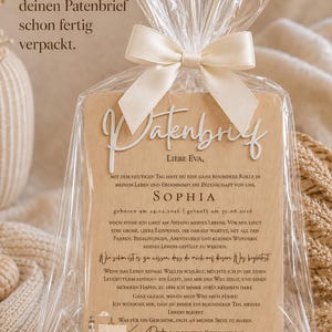 May include: A wooden plaque with the German text Patenbrief and a heartfelt message for a godparent, featuring a lighthouse illustration. The plaque is encased in a clear plastic bag tied with a cream-colored satin ribbon bow.