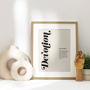 May include: A framed print with a beige background and black text that reads "Devotion". The text is stylized and vertical. The definition of the word is printed in black text below the word. The print is sitting on a white surface with two ceramic vases and a wooden hand.