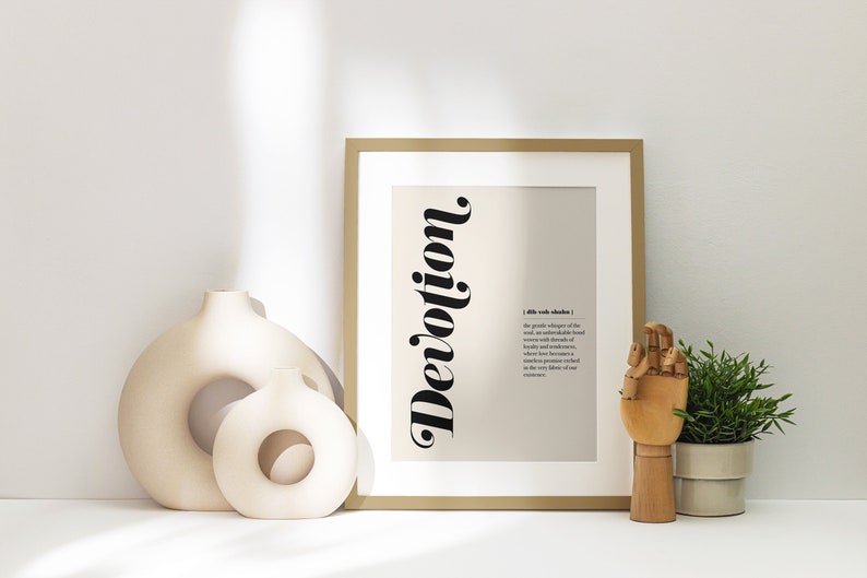 May include: A framed print with a beige background and black text that reads "Devotion". The text is stylized and vertical. The definition of the word is printed in black text below the word. The print is sitting on a white surface with two ceramic vases and a wooden hand.