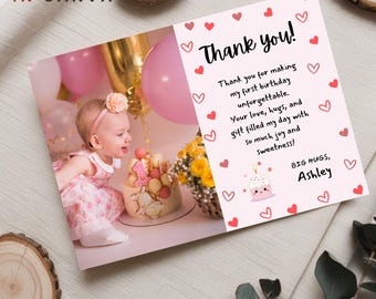 May include: A pink and white thank you card with a photo of a baby girl in a pink dress and a pink headband. The card has a pink and white cupcake with a candle and the text "Thank you!" in pink. The text on the card reads "Thank you for making my first birthday unforgettable. Your love, hugs, and gift filled my day with so much joy and sweetness! Big hugs, Ashley."