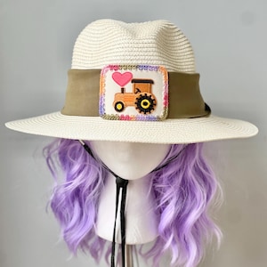 Peut inclure: Chapeau de soleil de couleur crème avec un large bord et un ruban marron. Le chapeau présente un écusson décoratif avec un tracteur et un cœur rose. Le chapeau est présenté sur une tête de mannequin avec des cheveux violets.