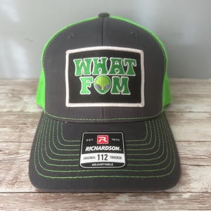 May include: A gray Richardson 112 trucker hat with a neon green mesh back and brim. The front features a patch with the words "WHAT FOM" in green, with a green alien graphic. The hat is adjustable.