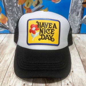 May include: Black and white trucker hat with a white front panel and a black brim. The hat features a yellow patch with the phrase "HAVE A NICE DAY" and a red flower graphic.