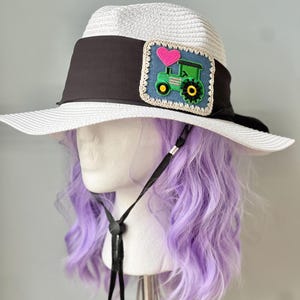 Peut inclure: Chapeau de soleil blanc à large bord avec un bandeau noir. Le chapeau est orné d'un écusson représentant un tracteur vert et un cœur rose. Une mentonnière noire est attachée. Le chapeau est présenté sur une tête de mannequin avec des cheveux violets.