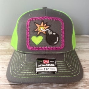 May include: Gray trucker hat with a neon green mesh back and brim. The front features an embroidered patch with a black bomb, a neon green heart, and an orange explosion. The patch is outlined in pink. The hat has the text "RICHARDSON ORIGINAL 112 TRUCKER ADJUSTABLE".